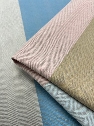 Close-up of folded fabric with a herringbone pattern from the Designer Cotton - Blipe - 155cm by Super Cheap Fabrics. The fabric features wide vertical stripes in pastel colors, including light pink, beige, blue, and grey, crafted using sustainable deadstock materials for eco-friendly fashion.