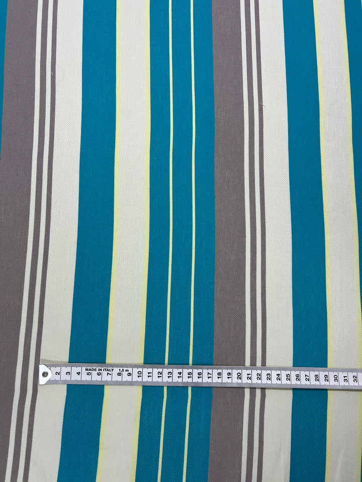 A striped fabric with vertical lines in various colors, including teal, gray, white, and yellow. Part of the Super Cheap Fabrics Designer Cotton - St.Tropez - 157cm range, this eco-friendly fashion piece features a measuring tape placed horizontally across the fabric, showing measurements in centimeters and inches.