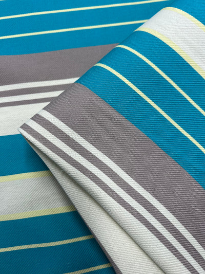 A close-up image of fabric from the Super Cheap Fabrics Designer Cotton - St.Tropez - 157cm Range featuring a pattern of horizontal stripes. The stripes vary in thickness and color, including shades of teal, gray, white, and yellow. The fabric is folded, showing layers of the pattern made from sustainable deadstock materials.