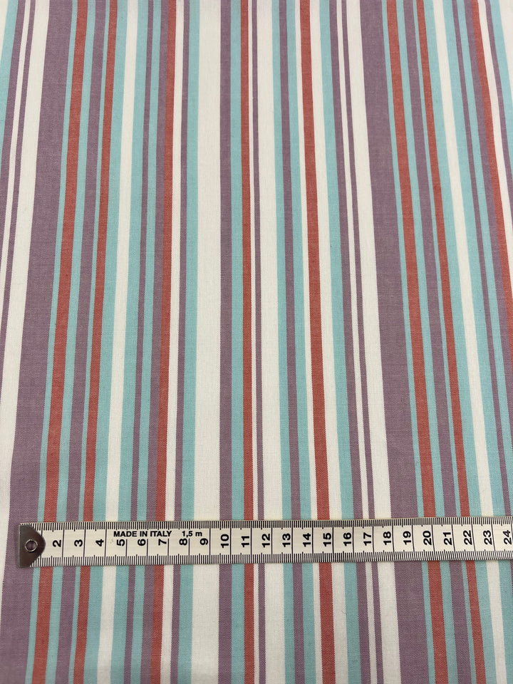 A measuring tape placed horizontally on a multicolored, striped Super Cheap Fabrics' Linen Cotton - Pulled Candy - 150cm fabric. The light-weight fabric features stripes in shades of purple, blue, orange, and white, while the measuring tape shows measurements in centimeters from 0 to 20.