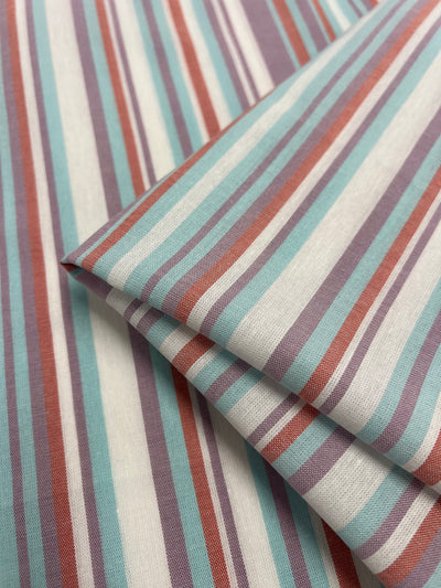Close-up of Super Cheap Fabrics' Linen Cotton - Pulled Candy - 150cm with vertical stripes in shades of turquoise, maroon, purple, and white. The natural fibers appear layered, with one piece folded over the other. The texture looks smooth and the colors are vibrant.