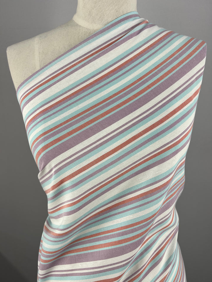A mannequin draped with Linen Cotton - Pulled Candy - 150cm by Super Cheap Fabrics, featuring a pattern of diagonal stripes in varying widths. Crafted from natural fibers combining linen and cotton, the stripes are in shades of light blue, light purple, white, and rusty pink against a plain gray background.