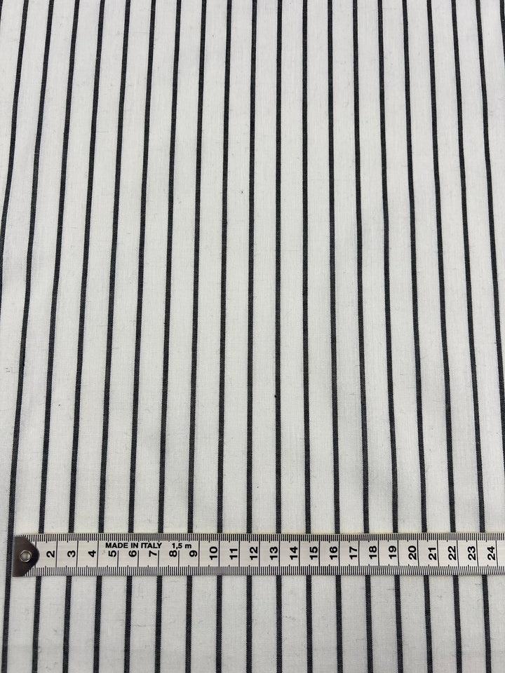 A close-up image of Super Cheap Fabrics' Linen Cotton - Pin Charcoal and White Stripe - 145cm with black vertical stripes. At the bottom of the image, there is a white measuring tape with black markings indicating centimeters and millimeters. The tape reads from 10 to 27 centimeters, showcasing the durable fabric's precision and quality.