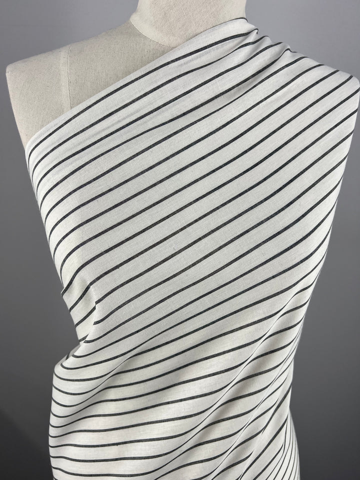 A mannequin is draped with a piece of durable fabric featuring a pattern of diagonal black stripes on a white background. Made from Linen Cotton - Pin Charcoal and White Stripe - 145cm by Super Cheap Fabrics, the fabric is wrapped around the mannequin's torso and over one shoulder. The background is plain gray.