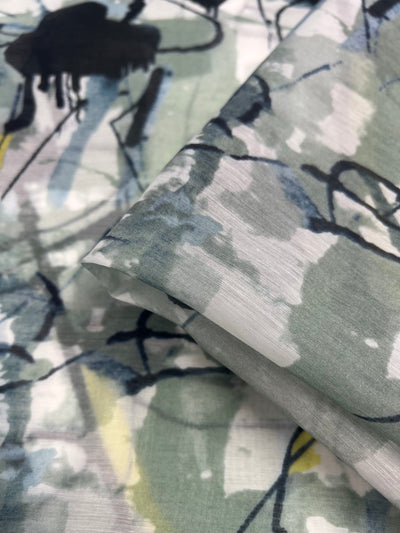 A close-up of a piece of lightweight, multi-colour fabric with an abstract pattern. The design features splashes and streaks of black, gray, green, blue, and yellow, creating a painterly effect. The fabric is smooth and slightly translucent, with part of it folded over. Ideal for children's clothing. Product Name: Designer Cotton - Branches - 150cm Brand Name: Super Cheap Fabrics