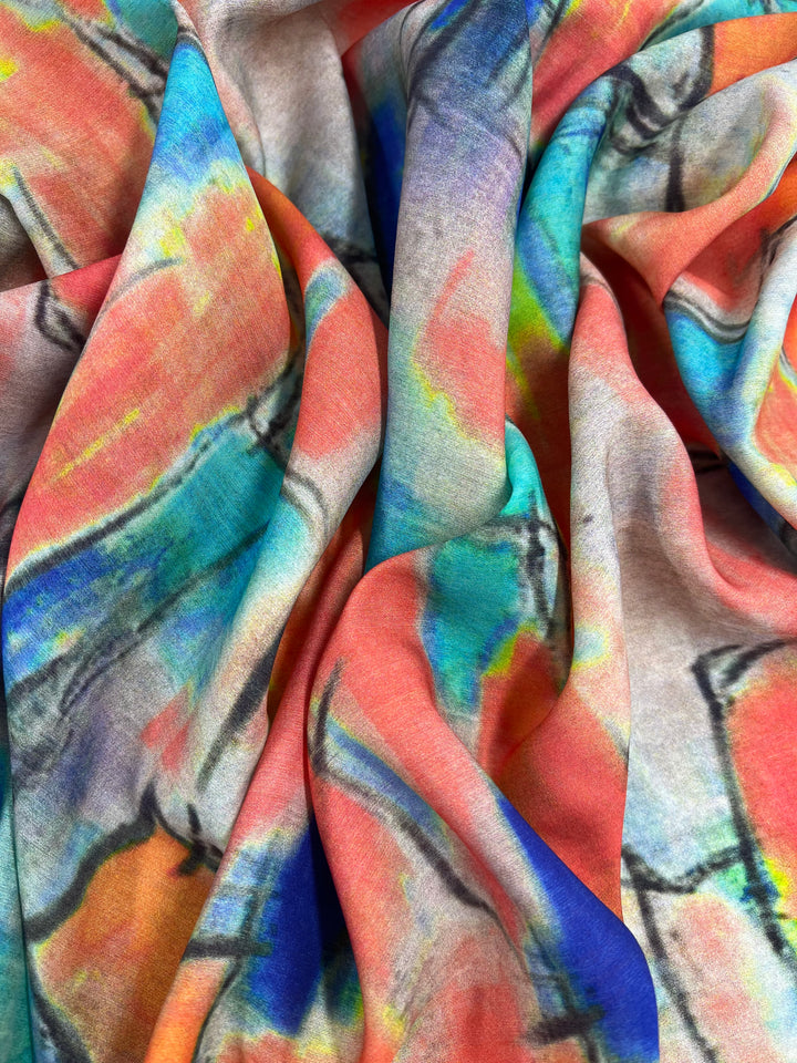 A close-up of colorful, crumpled fabric featuring a vibrant, abstract pattern with splashes of orange, blue, green, and yellow. Ideal for children's clothing or home decor, the light-weight fabric's folds create a dynamic and textured appearance. This is Designer Cotton - Infrared - 145cm by Super Cheap Fabrics.