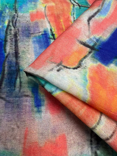 A close-up of Designer Cotton - Infrared - 145cm by Super Cheap Fabrics, ideal for home decor or children's clothing, featuring vibrant splotches and streaks of blue, red, orange, yellow, and green. The lightweight fabric is folded in the upper right corner, creating layers and a blend of hues with a watercolor-like appearance.