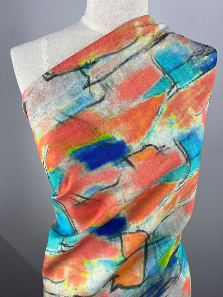 A mannequin is draped with a vibrant, abstractly patterned, lightweight fabric from Super Cheap Fabrics. The Designer Cotton - Infrared - 145cm features a blend of bright colors, including red, blue, turquoise, green, and yellow, along with black lines giving it a dynamic, artistic look. The background is a neutral gray.