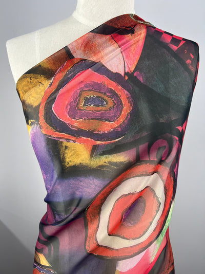 A mannequin is draped with brightly colored, abstract-patterned fabric from Super Cheap Fabrics. The Designer Chiffon - Geode - 150cm features bold swirls and circular shapes in vibrant red, pink, purple, and yellow hues against a dark background. Styled diagonally across the torso, this striking design evokes the elegance of fancy dresses.