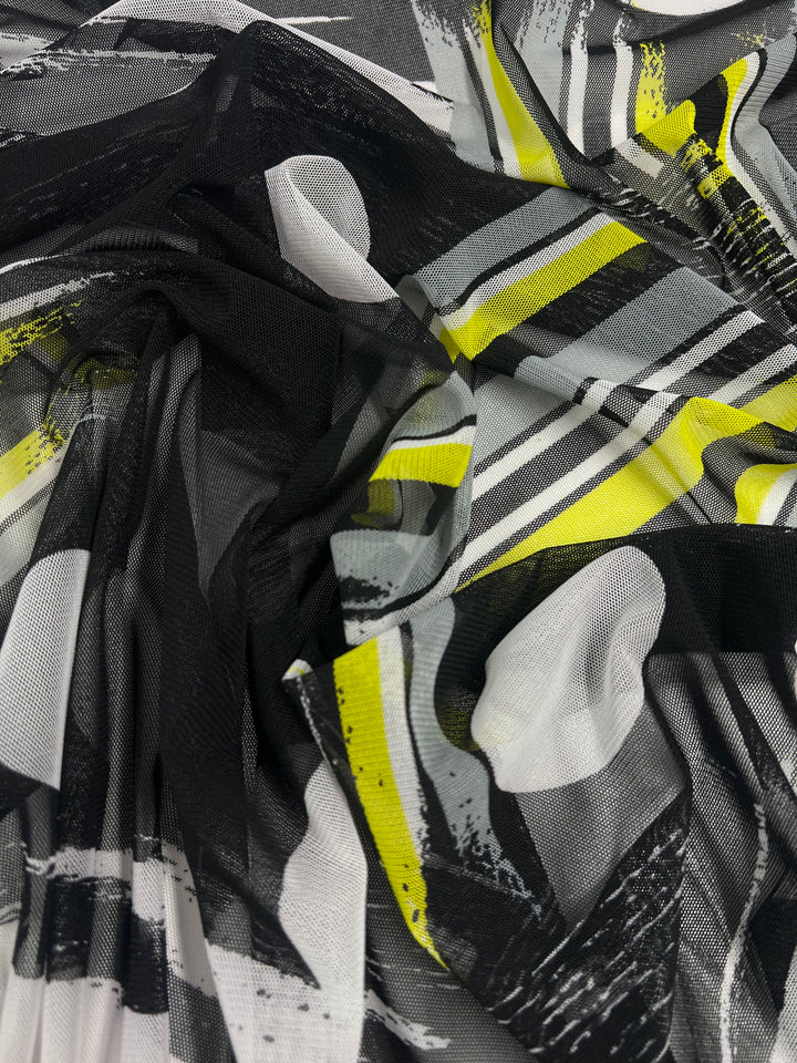 A piece of Super Cheap Fabrics Designer Mesh - Spot & Stripe - 150cm with an abstract pattern featuring black, white, gray, and yellow colors. The design includes bold, sweeping brushstrokes and overlapping shapes, giving the fabric a dynamic and modern appearance. Perfect for fancy dresses or wedding dresses, the fabric is slightly wrinkled and folded.