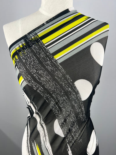 A mannequin draped with Designer Mesh - Spot & Stripe - 150cm by Super Cheap Fabrics featuring a modern geometric design suitable for fancy dresses. The fabric is black with a combination of white circles, yellow, gray, and white stripes, and abstract black and white patterns. The background is a neutral gray.