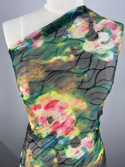 A mannequin draped with a vibrant fabric featuring a colorful floral pattern reminiscent of fancy dresses. The Designer Mesh - Neon Flora - 150cm from Super Cheap Fabrics has shades of green, pink, yellow, and black wavy lines, creating an abstract design. The background is a neutral gray color, highlighting the fabric's intricate details.