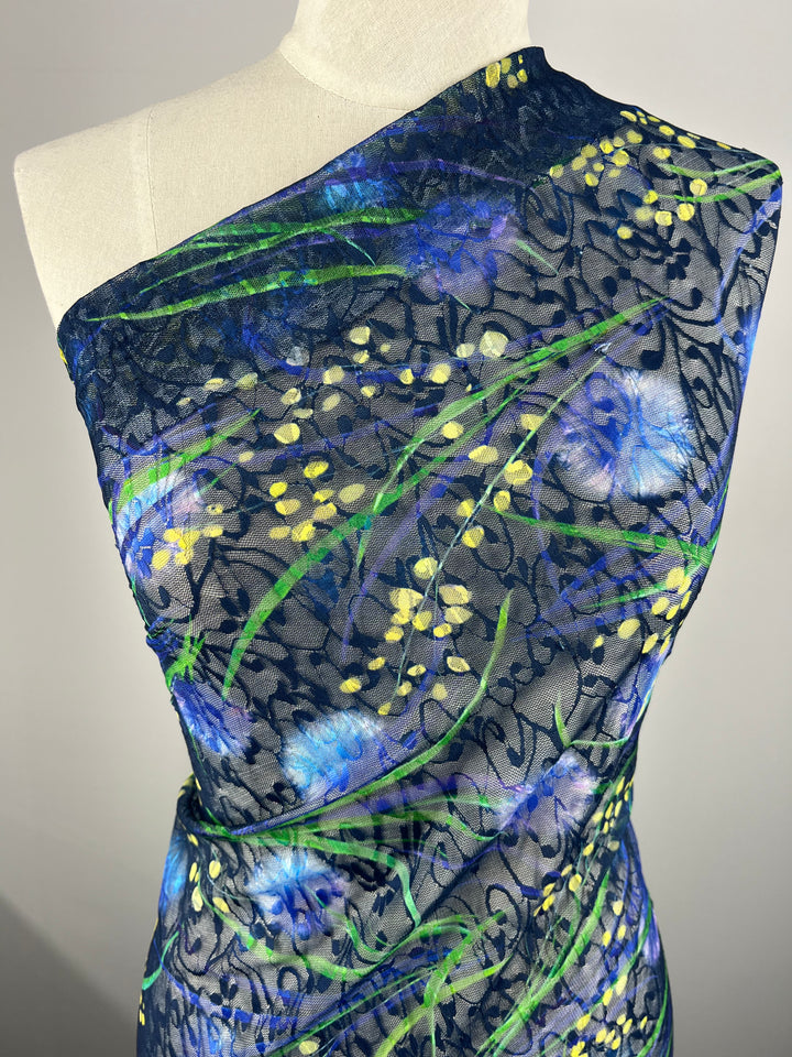 A mannequin draped in Super Cheap Fabrics' Designer Mesh - Lotus - 150cm, a sheer, one-shoulder fabric with a vibrant botanical design. The pattern features blue flowers, yellow buds, and green leaves on a dark mesh background. Made of 100% polyester, this lightweight fabric's intricate details are highlighted against the light backdrop.