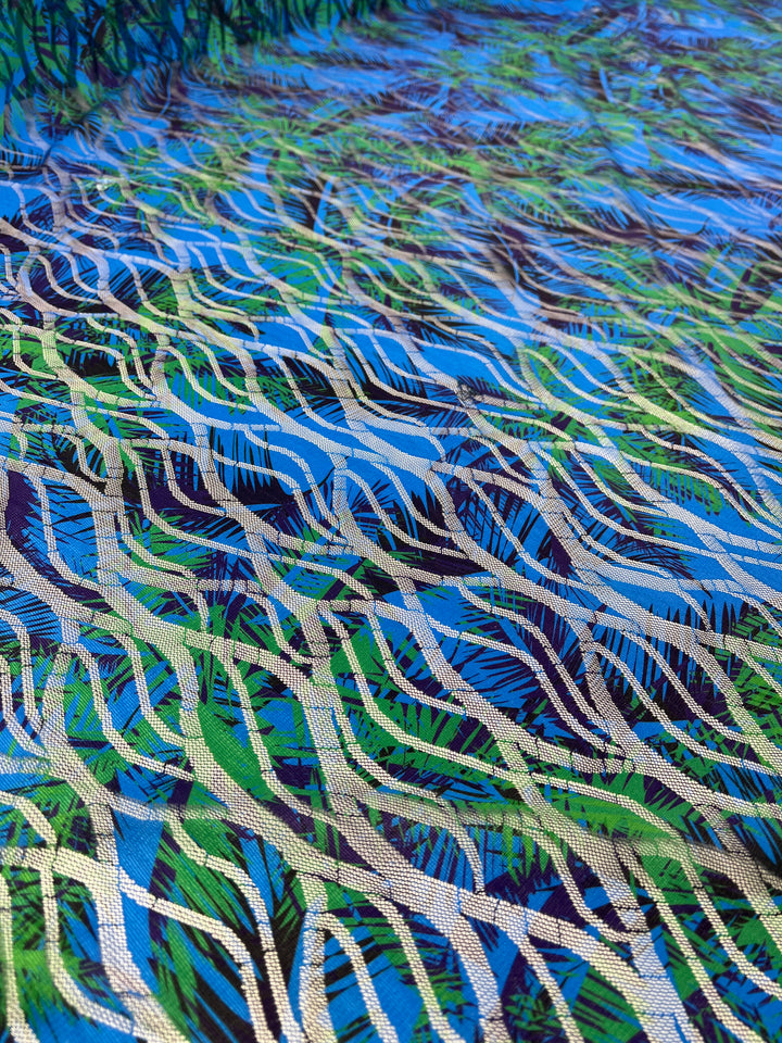 A vibrant, abstract pattern composed of intertwining green, blue, and beige wavy lines fills the image. Resembling flowing water or tall grass swaying, this multi-colour design on light weight fabric creates a dynamic and lively visual effect in the Super Cheap Fabrics' Designer Mesh - Palm Liquid - 150cm.