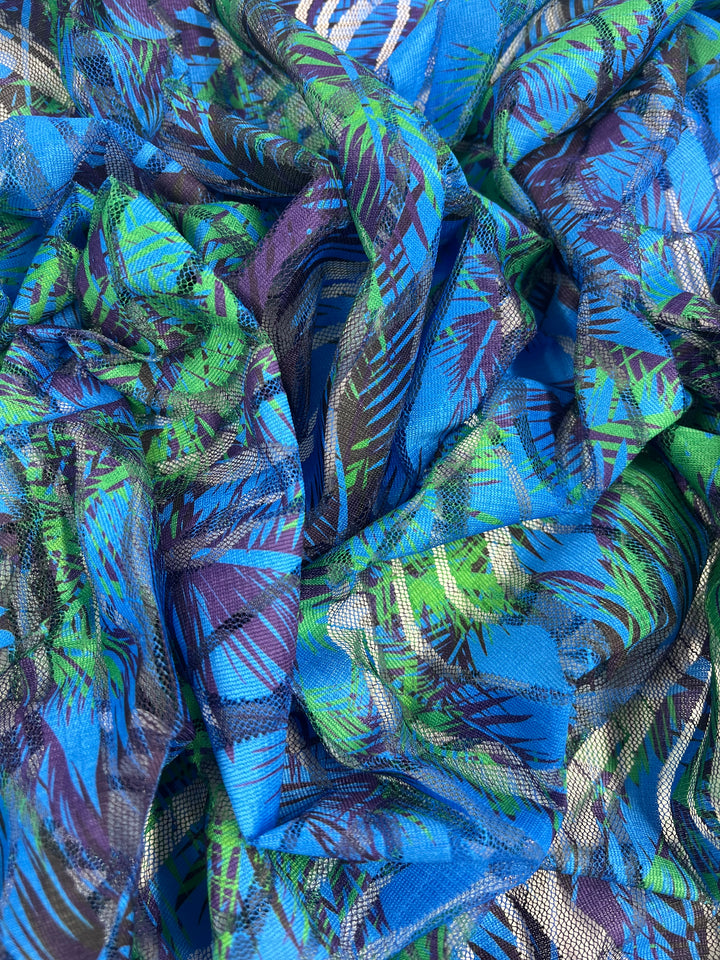 A close-up of a vibrant, multi-colored Designer Mesh - Palm Liquid - 150cm by Super Cheap Fabrics with an abstract pattern featuring shades of blue, green, and purple. The lightweight fabric appears crumpled with intricate, overlapping designs that include leaf-like shapes and striped elements.