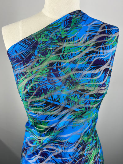 A mannequin is draped in a Designer Mesh - Palm Liquid - 150cm garment by Super Cheap Fabrics featuring a vibrant pattern of green, blue, and black abstract leaf designs on a bright blue background. The lightweight polyester fabric has a slight sheen, enhancing the multi-colour aesthetic.