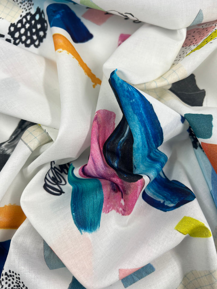 A close-up image of a white Designer Cotton - Design - 137cm fabric from Super Cheap Fabrics adorned with abstract, multicolored paint strokes. The design features vibrant colors including blue, pink, orange, yellow, and green, creating a dynamic and artistic pattern. The lightweight fabric is gently folded, adding a textured effect.