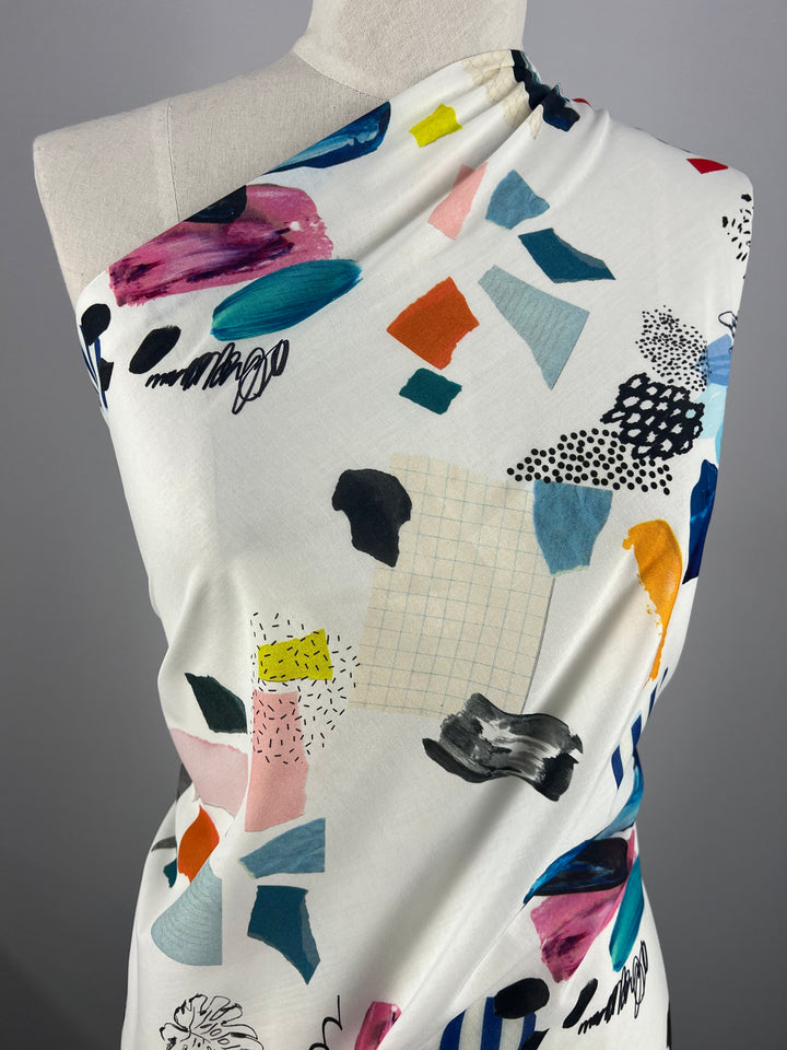 A close-up of a mannequin draped with Super Cheap Fabrics' Designer Cotton - Design - 137cm that features colorful abstract patterns. The multi-colour designs include various shapes in shades of blue, pink, orange, yellow, and black, with some grid lines and textured designs. The background is plain grey.