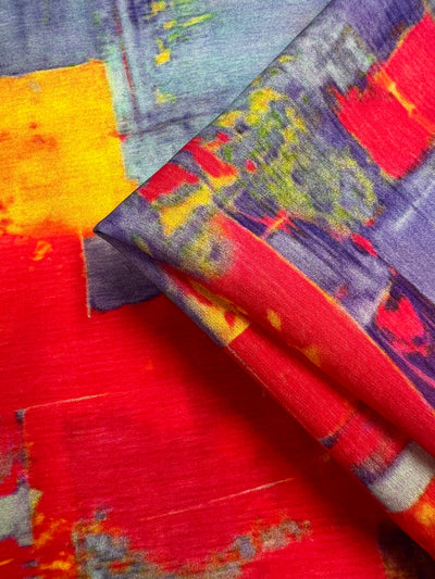 A close-up of a colorful, abstract cotton nylon fabric featuring bold geometric shapes in red, yellow, blue, and purple tones. The light weight Super Cheap Fabrics Designer Cotton - Fire and Ice - 145cm is neatly folded, displaying the vibrant pattern and texture.