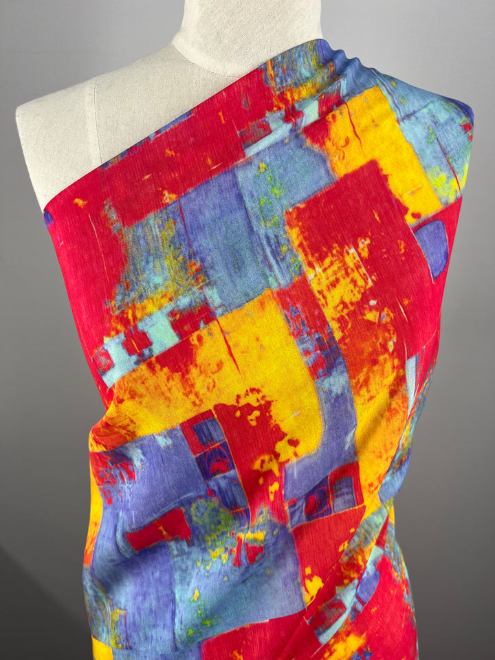 A mannequin is draped with Super Cheap Fabrics' Designer Cotton - Fire and Ice - 145cm, featuring a vibrant abstract pattern. The design includes bold patches of red, blue, yellow, and orange hues. The lightweight fabric is wrapped around the mannequin's shoulders, showcasing the dynamic and lively multi-colour print.