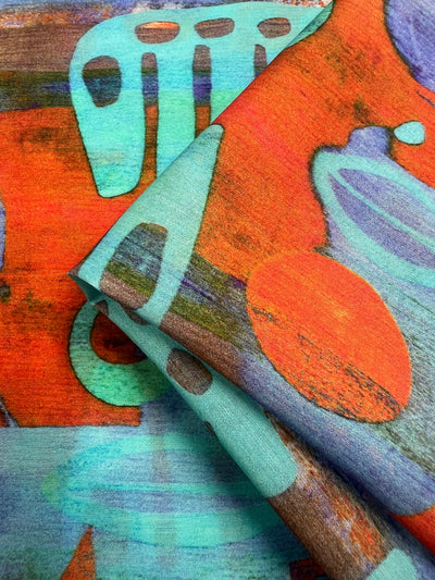 A close-up photo of colorful, folded fabric with abstract patterns. The Designer Cotton - Fossil - 150cm from Super Cheap Fabrics features vibrant shades of red, turquoise, purple, and orange with various shapes and lines, creating a visually dynamic and artistic design perfect for children's clothing.