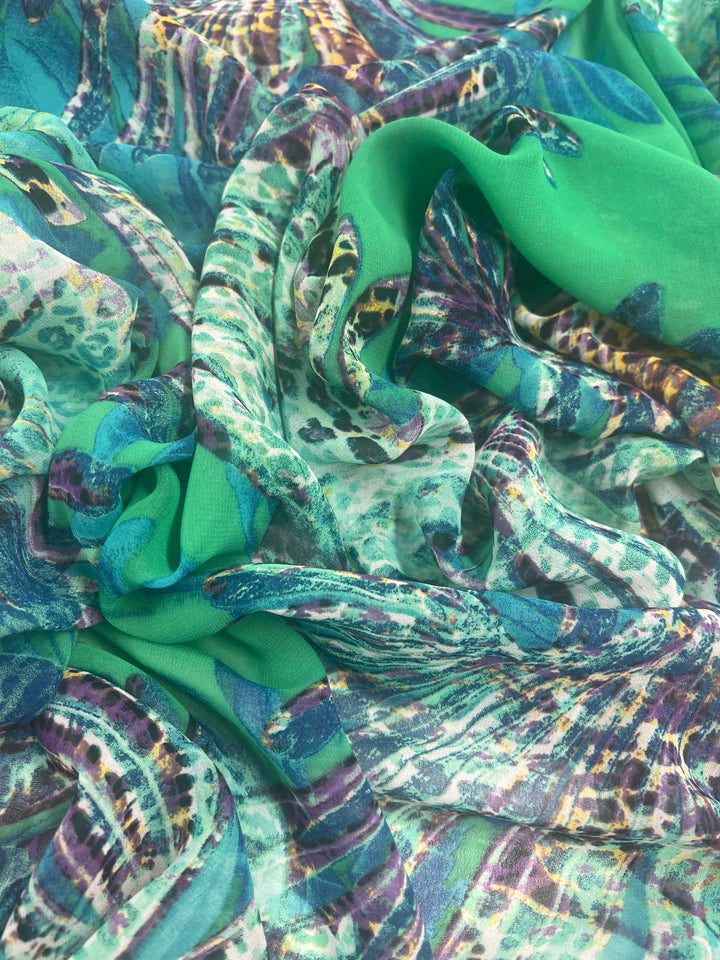 A close-up of a crumpled, colorful fabric featuring vivid green, turquoise, blue, and purple hues with abstract patterns and animal print elements. This Designer Chiffon - Leopard Palms - 150cm from Super Cheap Fabrics appears soft and semi-transparent with a mix of smooth and textured areas.