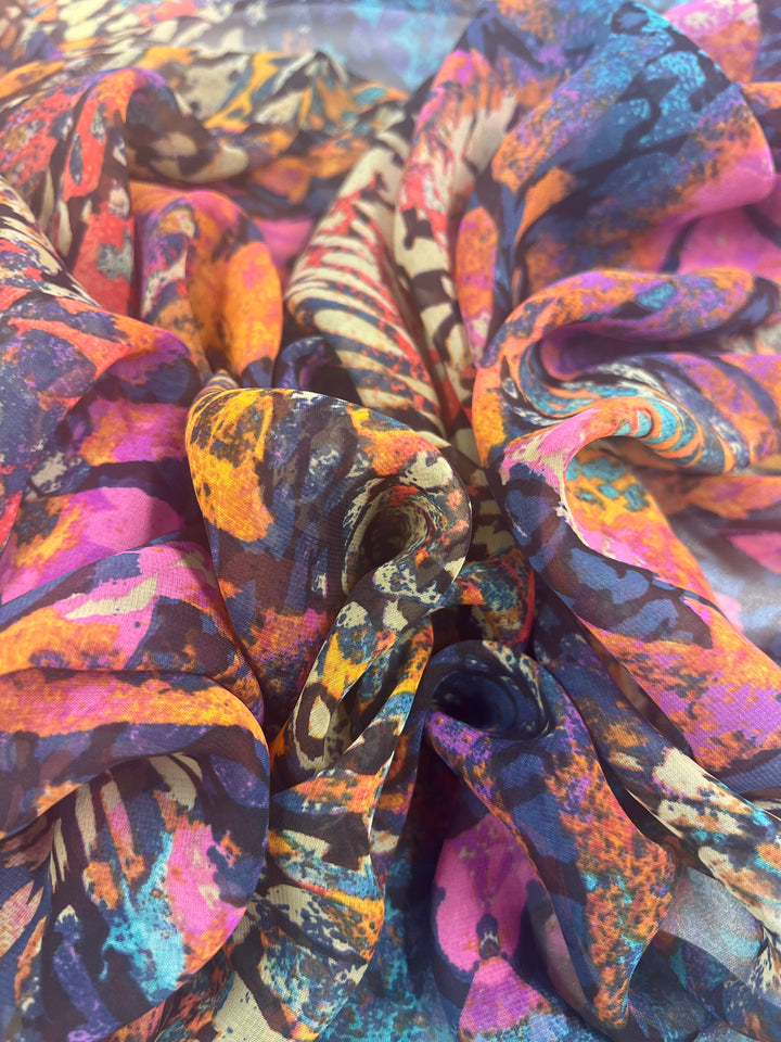 A close-up image of a colorful, abstract patterned fabric. The design features an array of vibrant colors including pink, orange, yellow, blue, and black, with a mix of swirling and folded textures. This extra lightweight fabric appears perfect for fancy dresses or stylish home décor. The product shown is Designer Chiffon - Tribadelic - 150cm by Super Cheap Fabrics.