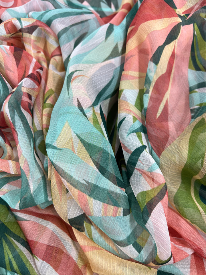 A close-up image of a colorful fabric with an abstract, tropical leaf pattern. The extra lightweight fabric features vibrant hues of pink, green, teal, and mustard yellow, creating a lively and intricate design. Perfect for fancy dresses or home décor projects, the material appears soft and slightly sheer. This is Super Cheap Fabrics' Designer Chiffon - Paradise - 150cm.
