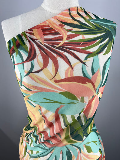 A mannequin is draped with colorful Designer Chiffon - Paradise - 150cm by Super Cheap Fabrics featuring a lively tropical leaf pattern. The fabric shows a mix of green, orange, yellow, and red hues arranged in a vibrant, overlapping design. The background is a simple, solid color, making it perfect for fancy dresses or home décor.