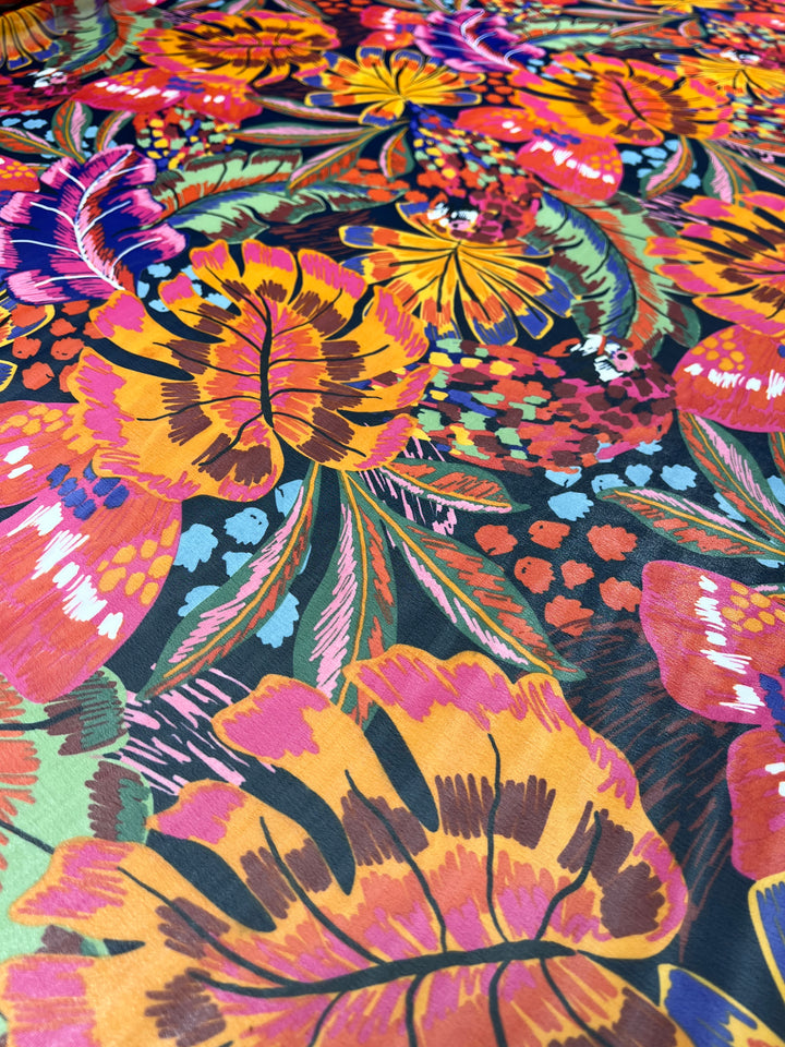 A vibrant, close-up image of colorful, tropical floral fabric often used in fancy dresses. The pattern features an array of large, overlapping flowers and leaves in shades of orange, pink, yellow, purple, and green on a dark background, creating a bold, lively, and intricate design. This is the Designer Chiffon - Wonderland - 150cm by Super Cheap Fabrics.