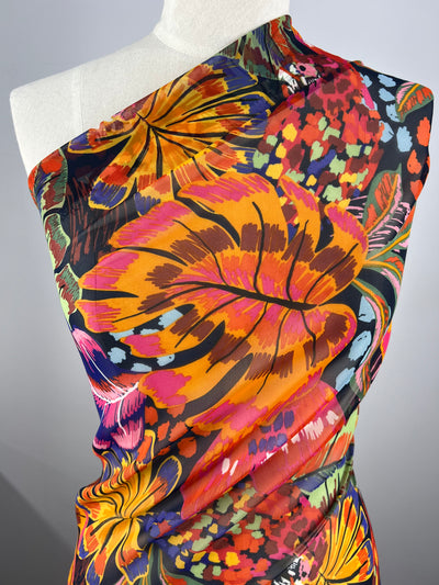 A mannequin draped in a colorful one-shoulder fabric featuring a vibrant floral pattern with large, bold flowers in hues of orange, pink, red, yellow, and blue against a dark background. The extra light weight Designer Chiffon - Wonderland - 150cm by Super Cheap Fabrics has subtle wrinkles and a flowing texture.