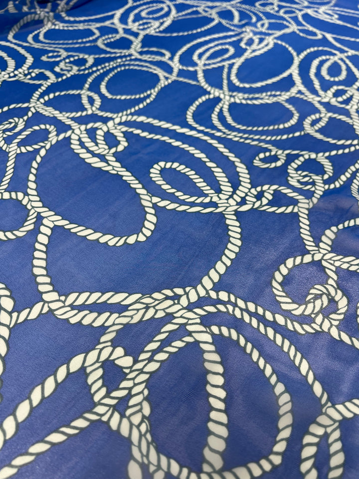A close-up view of Super Cheap Fabrics' Designer Chiffon - Ocean Rope - 150cm featuring intricate white rope patterns set against a rich blue background. The ropes are arranged in various overlapping loops and swirls, creating a complex and visually engaging design, perfect for adding an elegant touch to your home décor.
