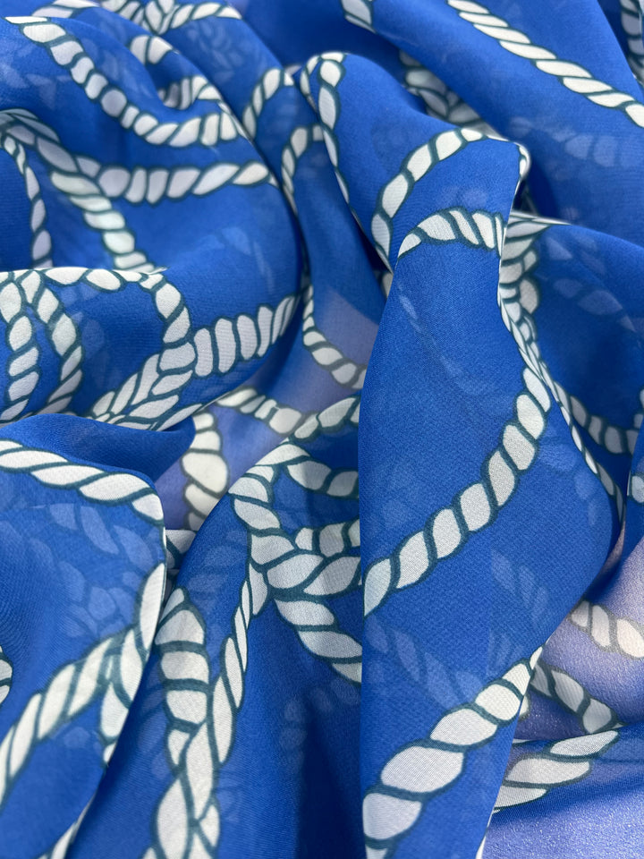 A close-up view of a blue fabric featuring a white twisted rope pattern, perfect for home décor. The Designer Chiffon - Ocean Rope - 150cm from Super Cheap Fabrics is gently crumpled, creating a textured and dynamic appearance. The rope design is evenly spaced, enhancing the nautical theme of the fabric.