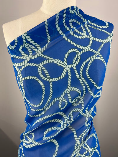 A mannequin draped with a sheer blue fabric featuring an intricate pattern of white, rope-like loops. The Designer Chiffon - Ocean Rope - 150cm fabric by Super Cheap Fabrics, perfect for fancy dresses or home décor, has a nautical theme and is displayed against a neutral background.