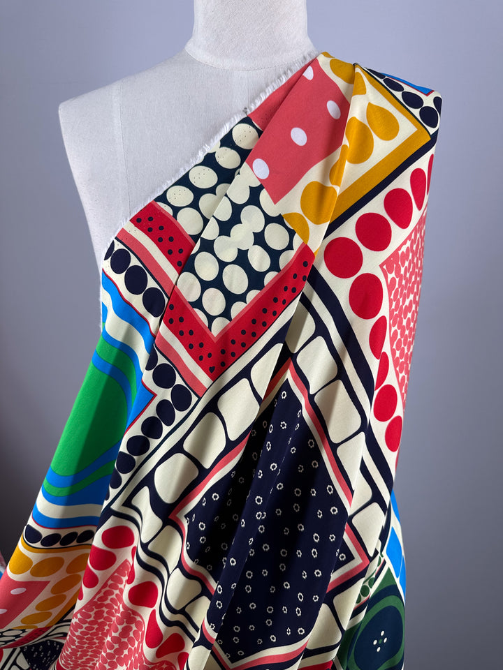 A white mannequin showcases Super Cheap Fabrics' Designer Rayon - Stile (145cm), featuring vibrant geometric shapes, polka dots, and bold red, green, yellow, navy, and cream prints. The lightweight rayon is draped diagonally across the torso.