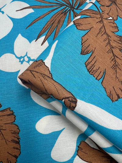 Close-up of tropical-themed, breathable fabric with a blue background. The design features large brown palm leaves and white hibiscus flowers. Made from **Super Cheap Fabrics Linen Cotton - Jasmine Breeze - 140cm**, the fabric is slightly folded, displaying a portion of the pattern prominently.