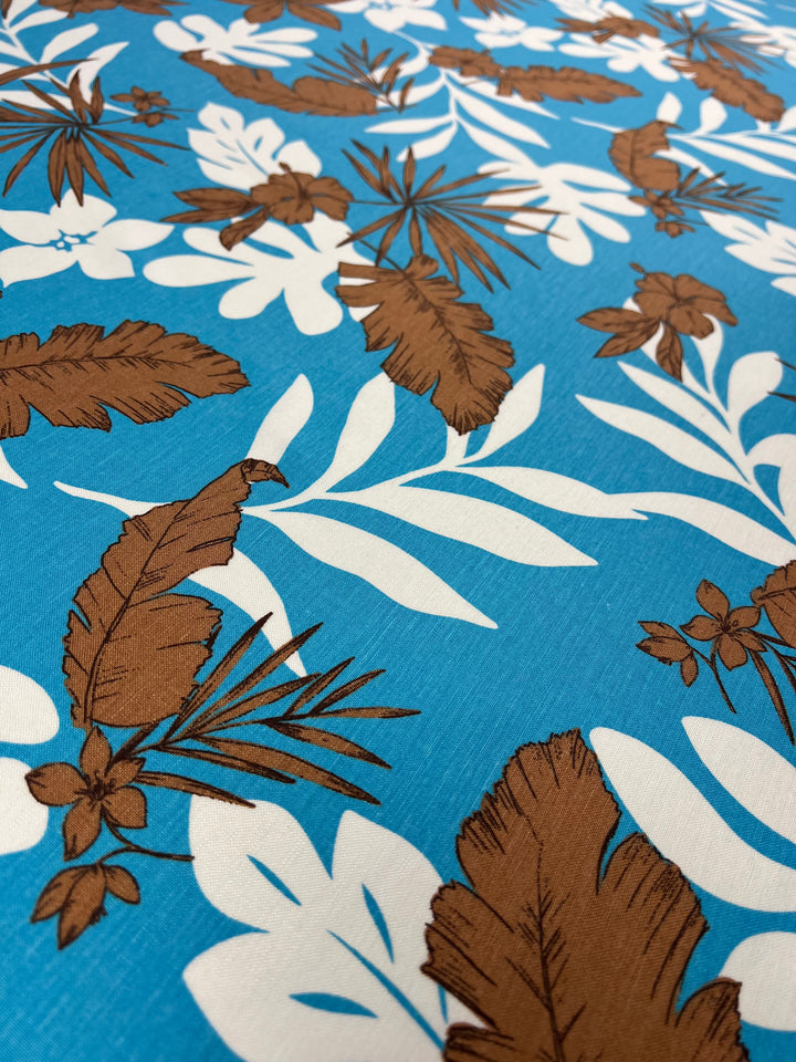 A close-up of a Linen Cotton - Jasmine Breeze - 140cm fabric with a vibrant tropical pattern. The design features large white leaves and brown palm fronds intermixed with small brown flowers on a bright blue background. This breathable fabric from Super Cheap Fabrics, made from natural fibers, has a textured appearance.