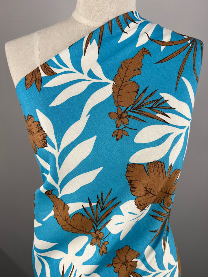 A blue fabric is draped over a dress form. The Linen Cotton - Jasmine Breeze - 140cm from Super Cheap Fabrics features a tropical print with large white leaves and brown hibiscus flowers, creating a vibrant, summery pattern against the bright blue background. Its breathable fabric ensures comfort during warm weather.