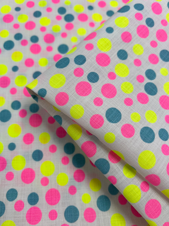 A close-up of a folded Linen Cotton - Spotty - 150cm from Super Cheap Fabrics with a playful, colorful polka dot pattern. The dots are in shades of neon pink, yellow, and dark teal on a white background, creating a vibrant and cheerful design. This breathable fabric made from natural fibers ensures comfort and style.