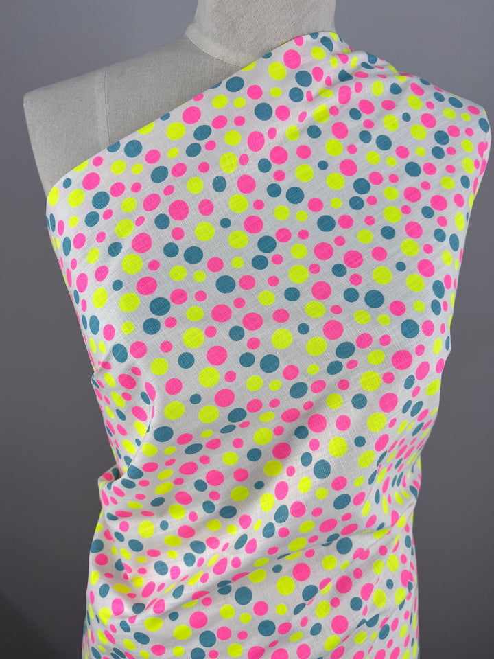 A mannequin draped in Super Cheap Fabrics' Linen Cotton - Spotty - 150cm adorned with a colorful polka dot pattern of neon pink, yellow, and blue dots posed against a plain gray background. Crafted from breathable Linen Cotton natural fibers, the fabric extends elegantly from the mannequin's shoulder downwards.