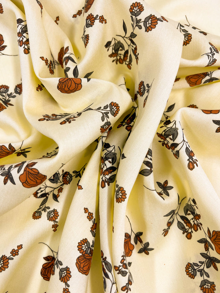 A close-up of a cream-colored Printed Rayon - Calico Flor - 145cm with a floral print from Super Cheap Fabrics. The pattern includes small, delicate flowers and leaves in shades of orange and black, scattered across the fabric which is softly gathered and draped, creating elegant folds and textures.