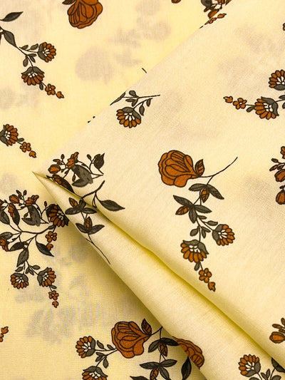 A close-up image of **Super Cheap Fabrics' Printed Rayon - Calico Flor - 145cm** features a scattered pattern of small brown and orange flowers with green leaves. Some parts of the fabric are folded, showing the design from different angles. The floral pattern creates a delicate and whimsical look, making it a versatile choice.