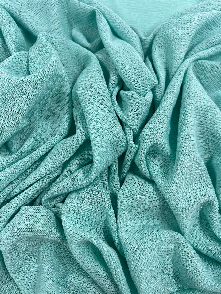 A close-up of a piece of the Lightweight Slub Jersey - Aqua - 150cm from Super Cheap Fabrics with a mesh-like texture, arranged in soft, flowing folds. The fabric appears lightweight and drapey, creating gentle curves and intricate patterns. This unique design offers maximum comfort for any stylish outfit.