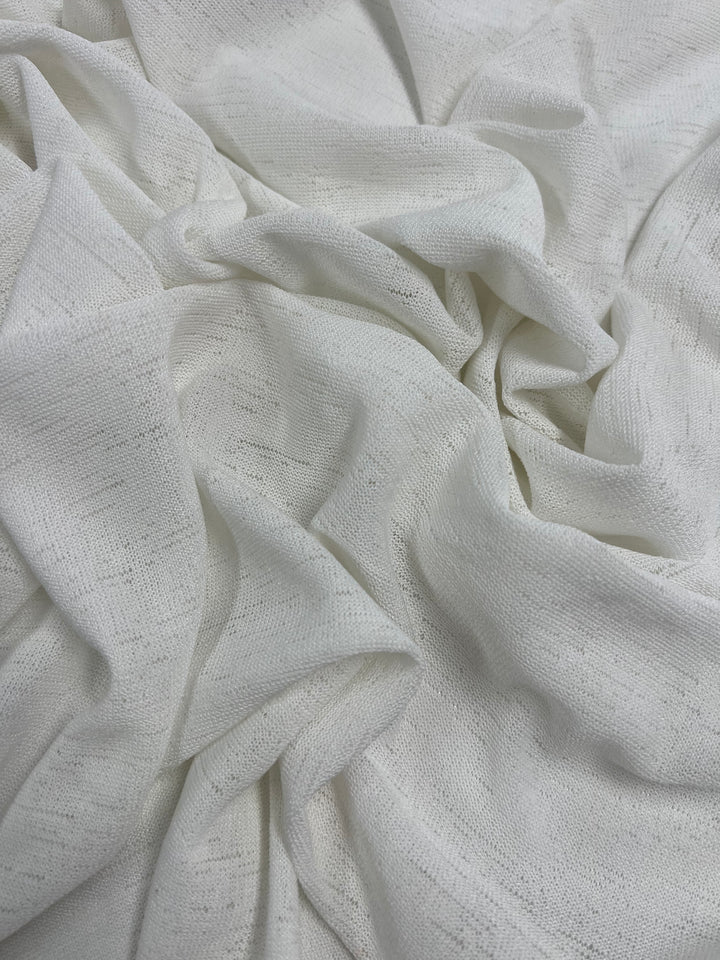 A close-up of crumpled, white fabric with a slightly textured surface. The Lightweight Slub Jersey - Ivory - 150cm from Super Cheap Fabrics appears soft and lightweight, creating subtle shadows and folds across its stretchable surface. The image showcases the intricate details and weave pattern of this versatile clothing fabric.