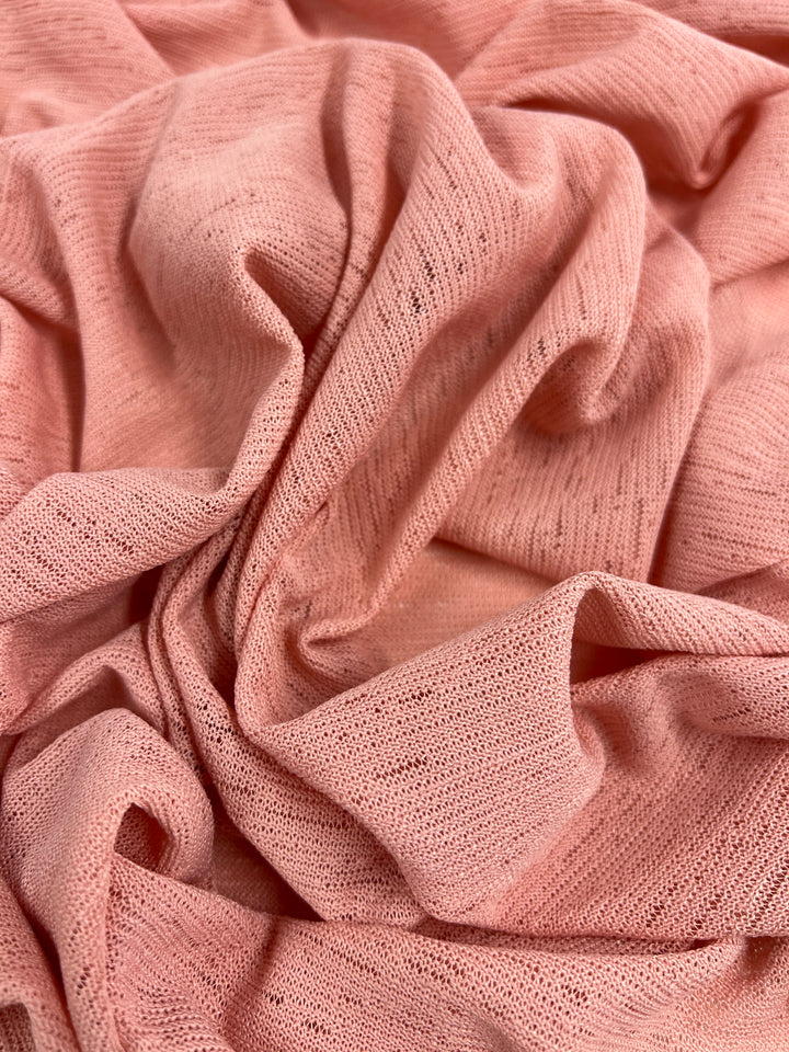 A close-up view of a soft, light pink fabric with a slightly crinkled texture. The 100% Polyester material appears stretchy and lightweight, exhibiting a delicate and airy feel with subtle variations in the textured knit pattern. This is the Lightweight Slub Jersey - Blossom - 150cm by Super Cheap Fabrics.