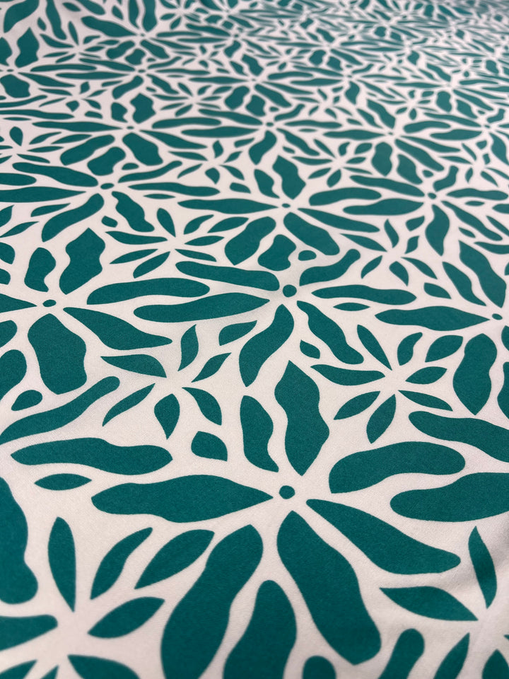 A close-up view of a Super Cheap Fabrics Designer Rayon - Ceramic - 140cm fabric featuring a repetitive green floral design with petal-like shapes on a white background. The vibrant prints create a visually striking and symmetrical layout, perfect for versatile fabric projects.