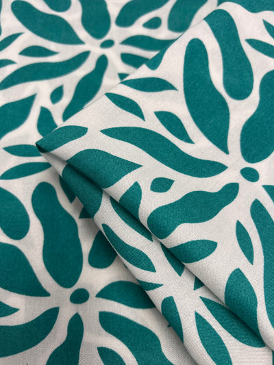 A close-up image of a folded Super Cheap Fabrics Designer Rayon - Ceramic - 140cm fabric featuring a bold turquoise and white abstract leaf pattern. The versatile fabric is neatly stacked, showcasing the intricate and vibrant design. The background consists of similar patterns, creating a cohesive and visually appealing look.