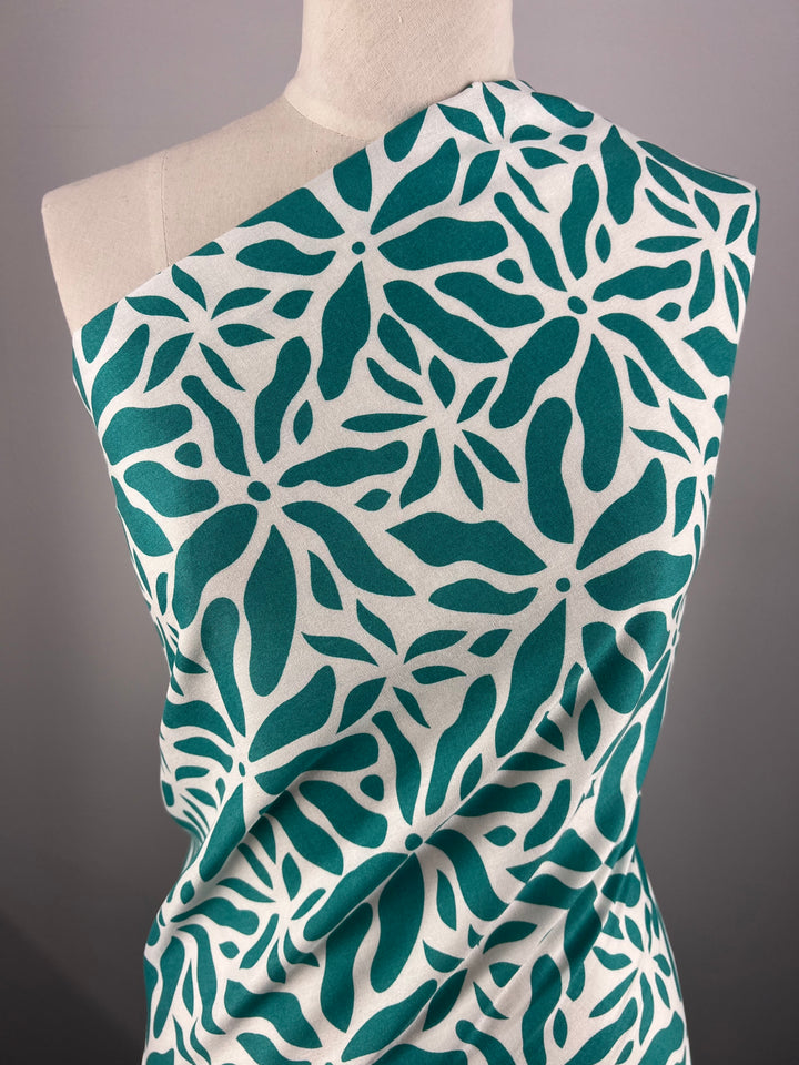 A mannequin is draped with Super Cheap Fabrics' Designer Rayon - Ceramic - 140cm featuring a green and white abstract leafy pattern. The vibrant print is wrapped in a one-shoulder style, covering the upper torso, against a neutral gray background.