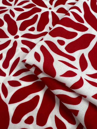 Close-up of Super Cheap Fabrics Designer Rayon - Scarlet - 140cm featuring an abstract pattern. The design consists of irregular red shapes resembling leaves or petals against a white background, creating a vibrant and dynamic visual effect. The versatile fabric is partially folded.