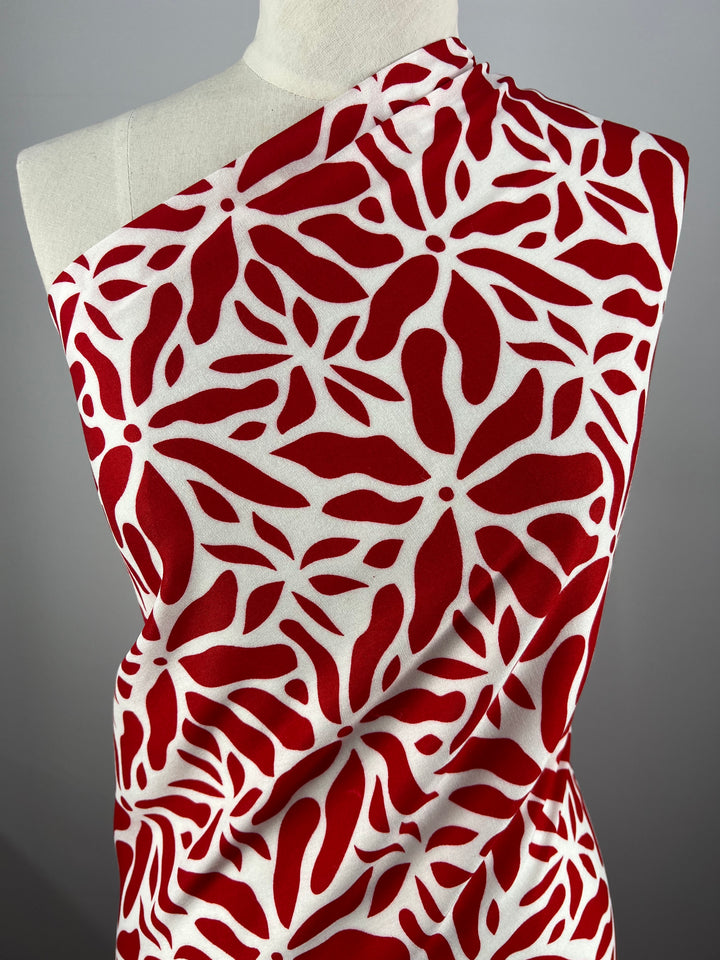 A mannequin displays a one-shoulder garment crafted from Super Cheap Fabrics' Designer Rayon - Scarlet - 140cm, featuring a bold red and white abstract leaf pattern. Set against a neutral gray backdrop, the vibrant print and versatile fabric create a striking visual contrast.